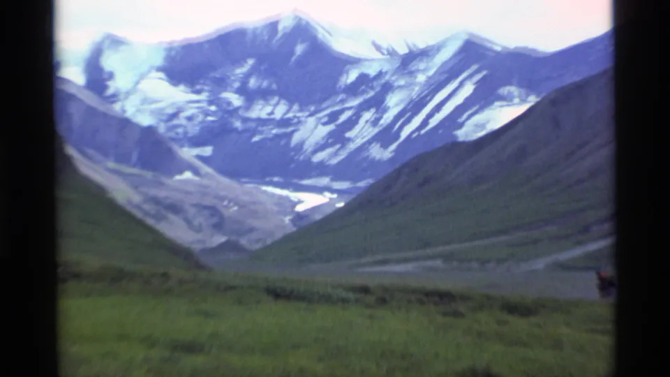 1977 Alaska 8mm frame looking deeper into an alpine valley, with snow-covered mountains and a pale glacier or river course below.