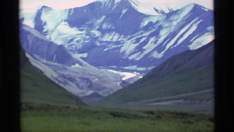 1977 Alaska home-movie frame centered on snow-capped peaks above a broad valley with a pale glacier or river channel.