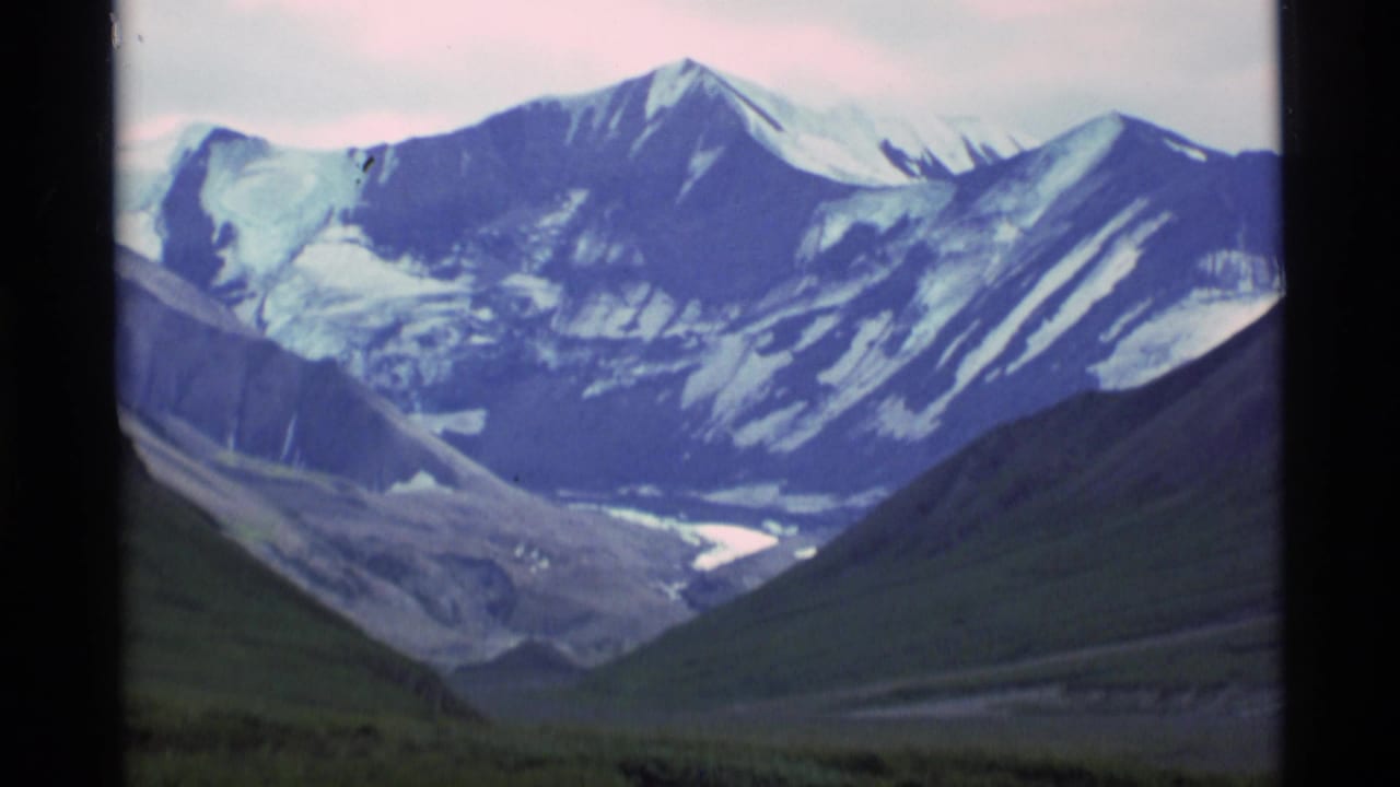 1977 Alaska 8mm frame showing snow-capped peaks and a wide mountain valley with a pale sky.