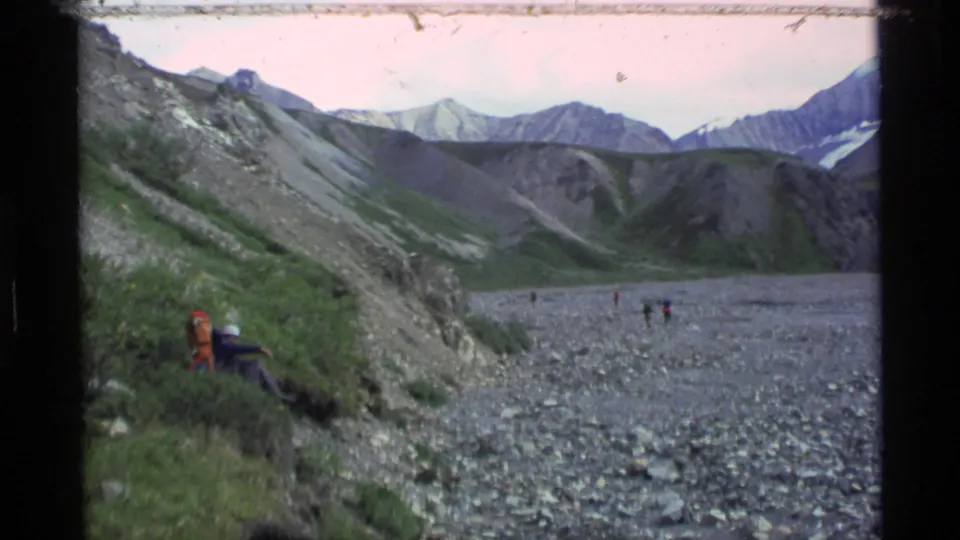 1977 Alaska home-movie frame with distant hikers and a backpack near a rocky riverbed in a mountain valley.
