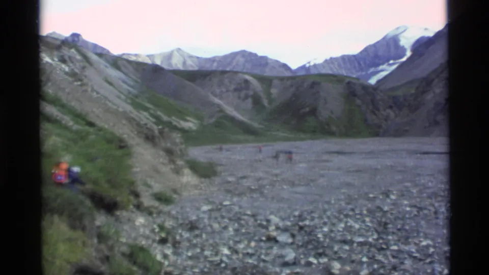 1977 Alaska 8mm frame showing backpacked hikers near a rocky riverbed with mountains beyond.