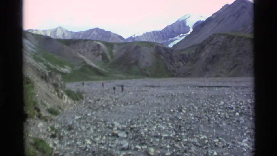 1977 Alaska home-movie frame with small hikers crossing a broad rocky valley beneath snow-capped peaks.