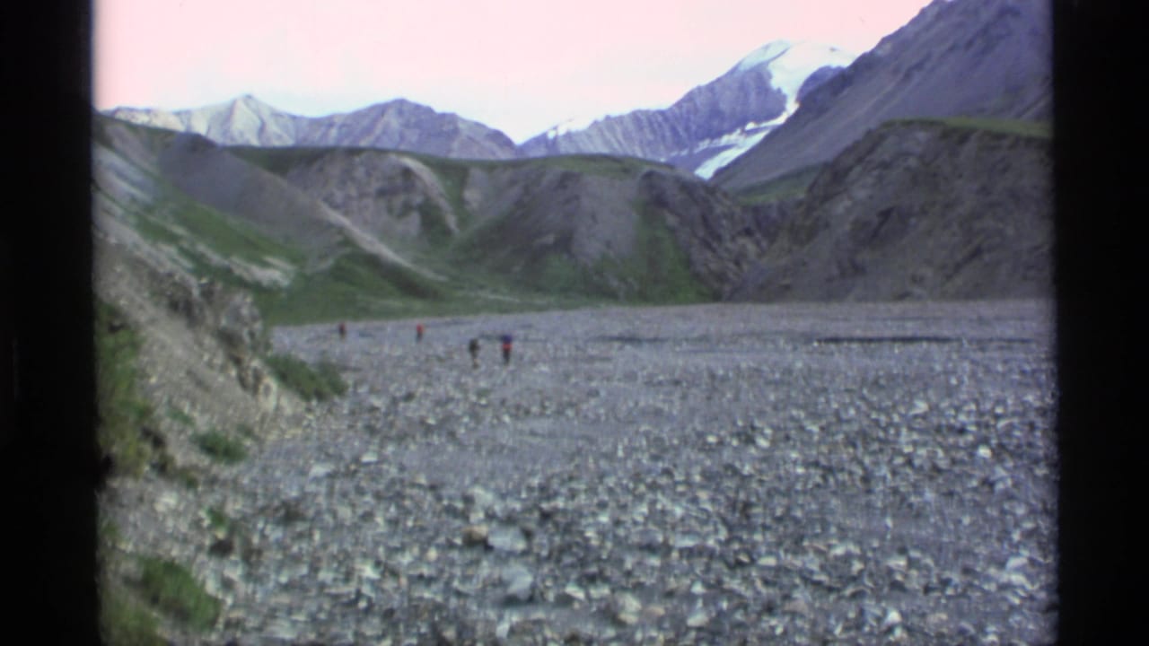 1977 Alaska 8mm frame showing a broad rocky river valley, small distant hikers, steep slopes, and snow-patched mountains under a pale sky.