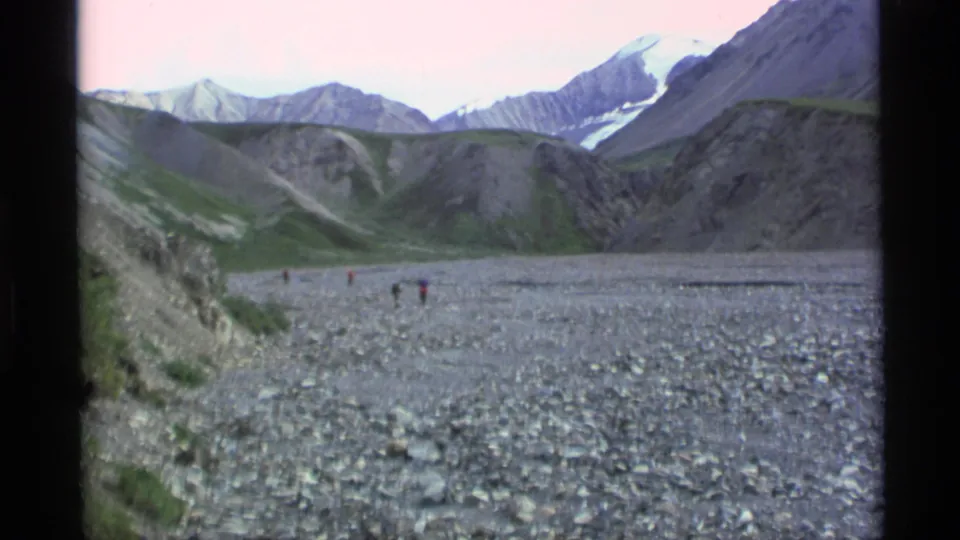 1977 Alaska 8mm frame showing a broad rocky river valley, small distant hikers, steep slopes, and snow-patched mountains under a pale sky.