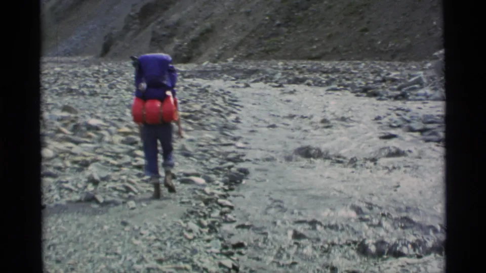 1977 Alaska 8mm frame following a backpacker with a blue pack and red sleeping gear across a stony riverbed in a mountain valley.
