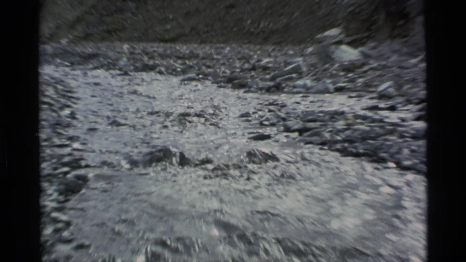 Close 8mm view of shallow water flowing through a rocky streambed, with blurred stones and steep terrain in the background.