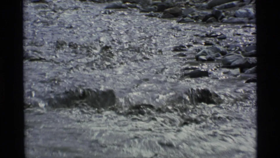 1977 Alaska 8mm frame showing shallow, fast-moving water running over rocks within a rugged mountain riverbed.