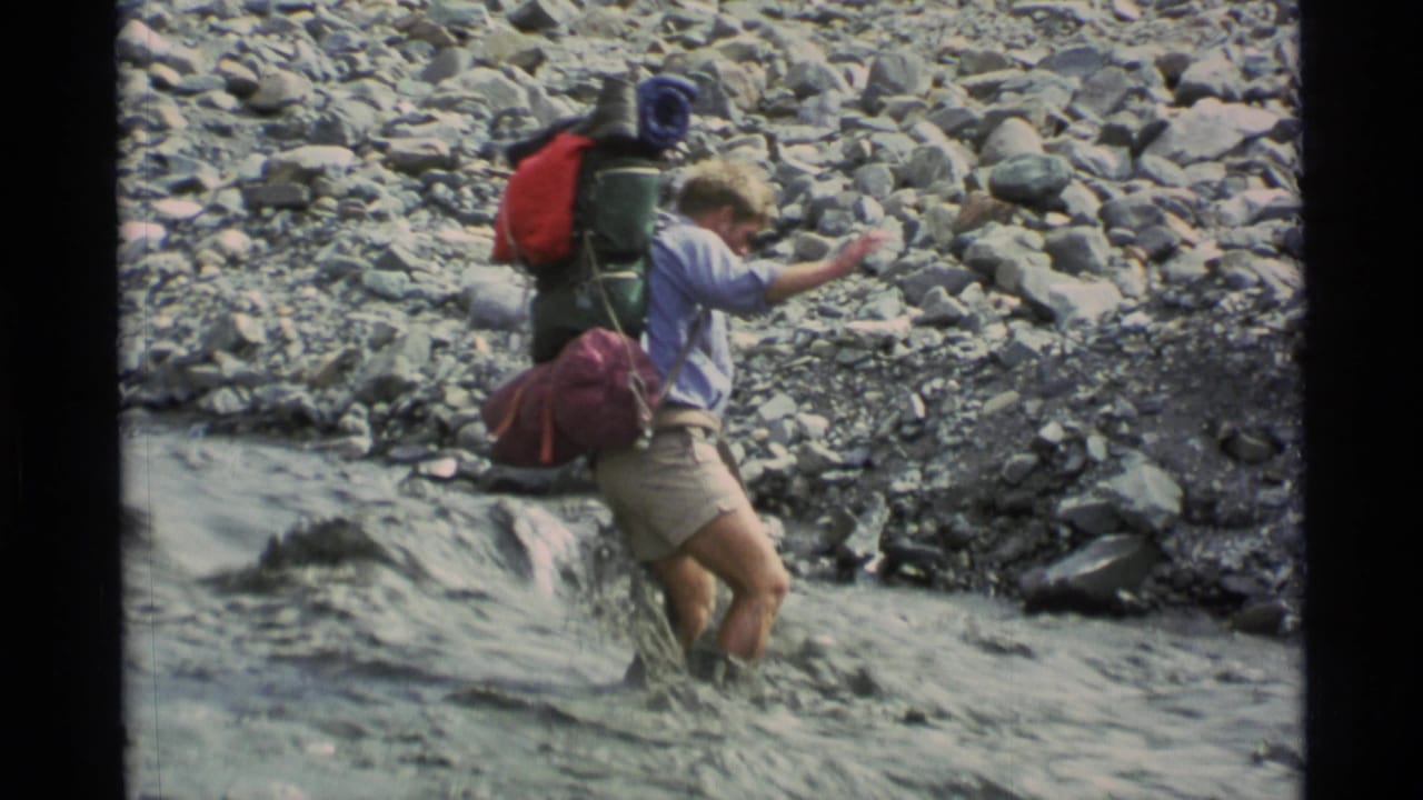 Hiker with a large backpack steps through a shallow rocky stream.