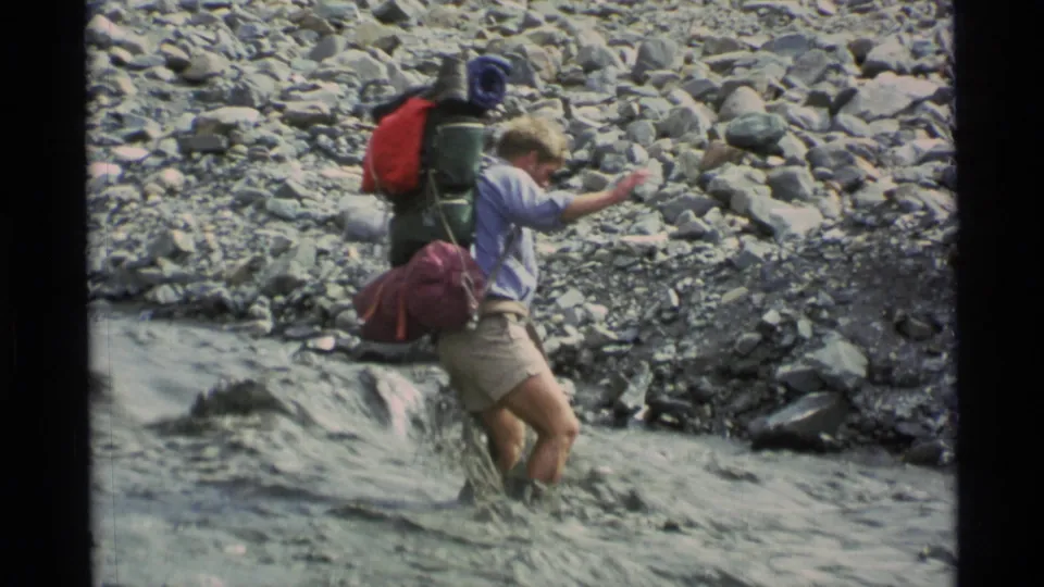 A backpacker in Alaska, 1977, steps carefully through shallow moving water beside a gray rocky bank.