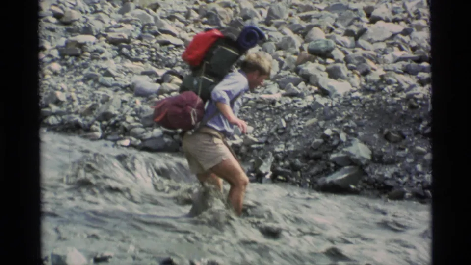 The hiker continues across the Alaska stream with red and green pack gear and a rolled blue item visible above his shoulders.