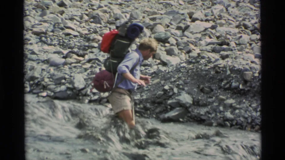 A backpacker in light shirt and shorts looks down toward the stream as water splashes around his legs in Alaska, 1977.