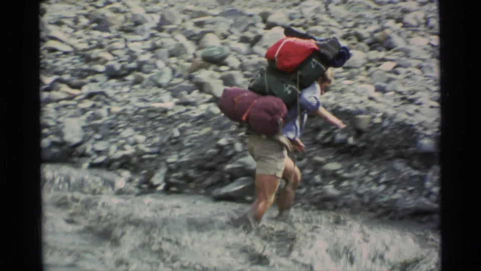 Near the end of the crossing, the loaded hiker leans forward toward the rocky bank as water rushes around his legs.