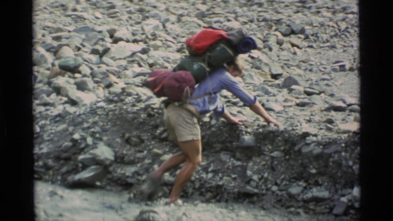 In 1977 Alaska home-movie footage, a hiker with a large backpack reaches across a rocky creek bed while crossing shallow water.