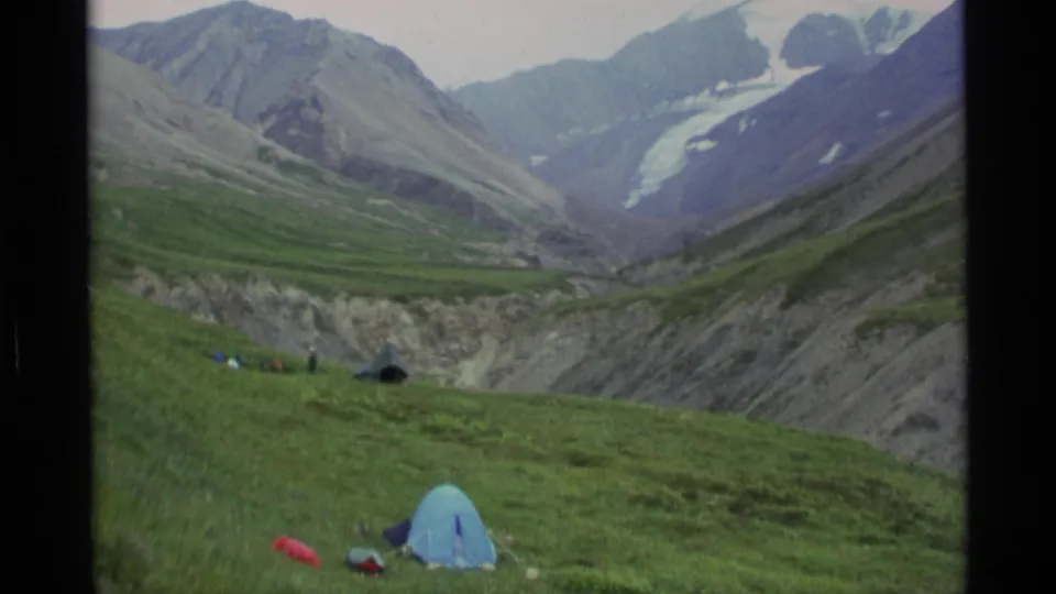 A wide Alaska mountain valley view shows tents pitched on green tundra-like grass with steep ridges and distant glacier-like snowfields.