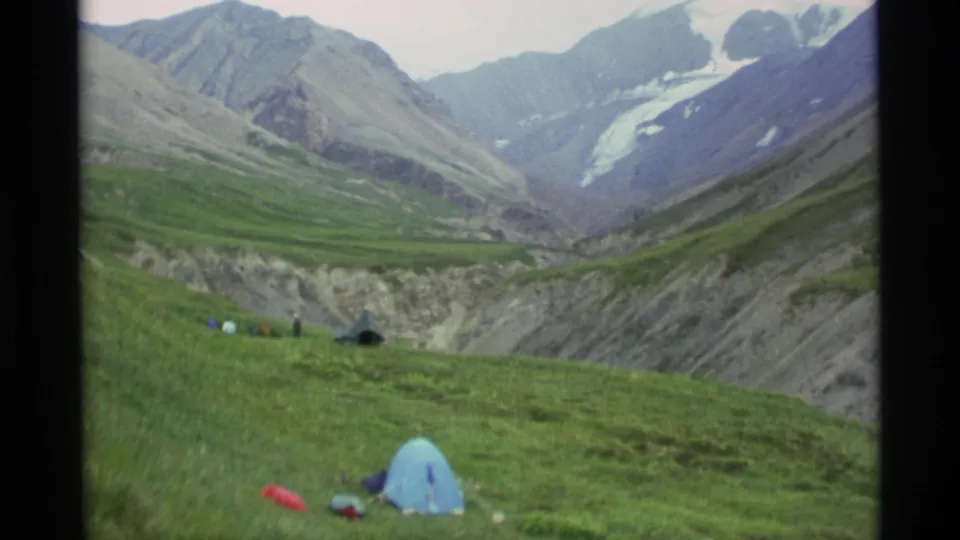 The home-movie frame shows a remote Alaska campsite with small tents and distant people on a green slope under a broad mountain valley.