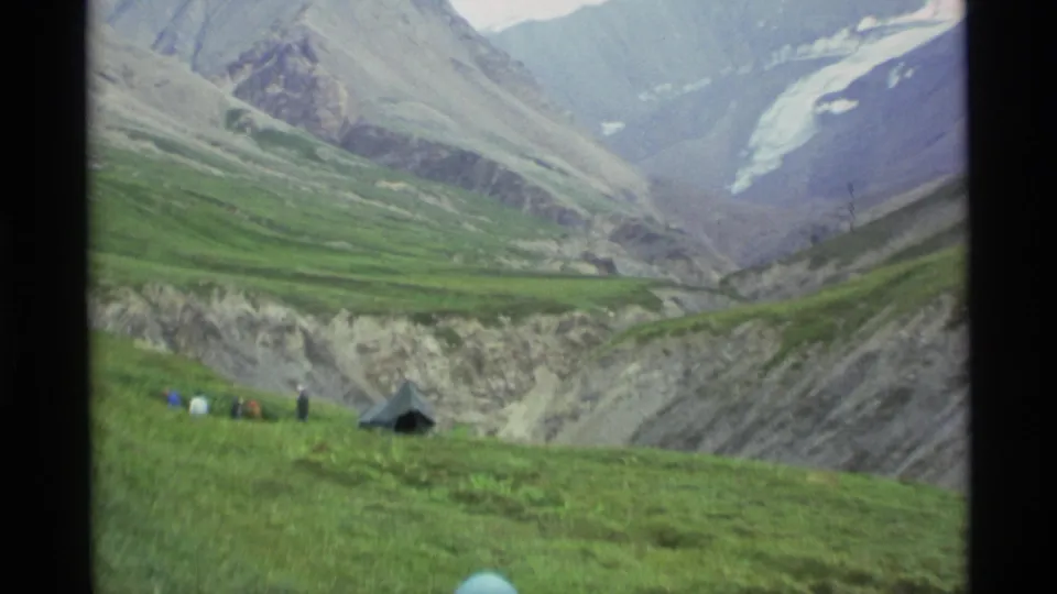 A dark tent and small distant campers stand on a grassy Alaska slope, with steep gray mountains and glacier-like snowfields beyond.