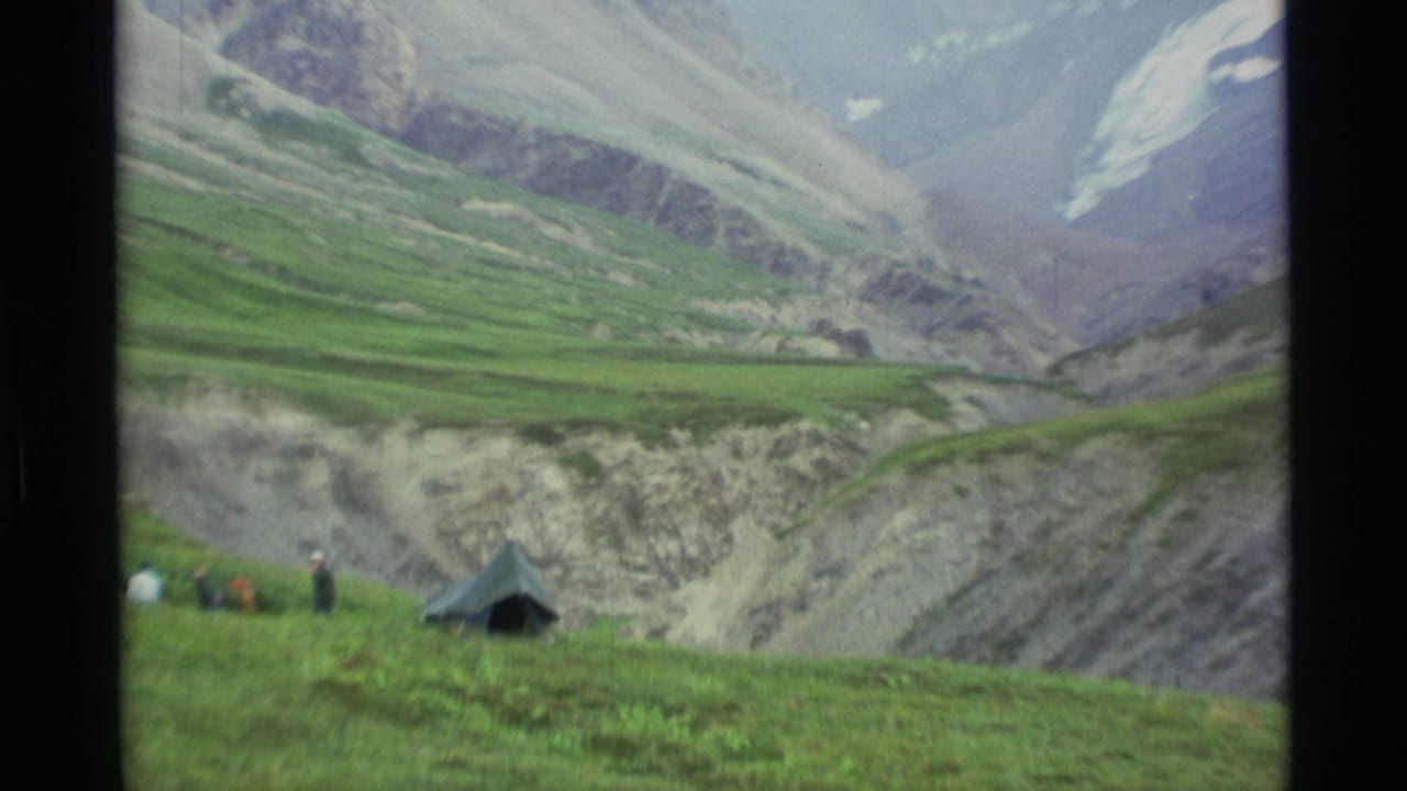1977 Alaska 8mm frame showing a green tent and distant people on a grassy slope beneath layered rocky mountains.