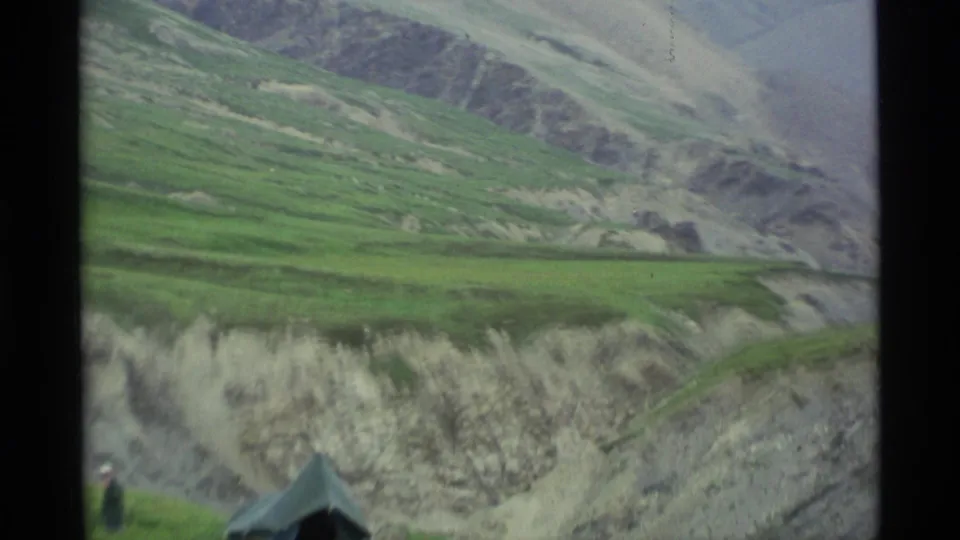 1977 Alaska 8mm view with the tent partly cropped, a distant person, green hillside, and eroded mountain terrain.