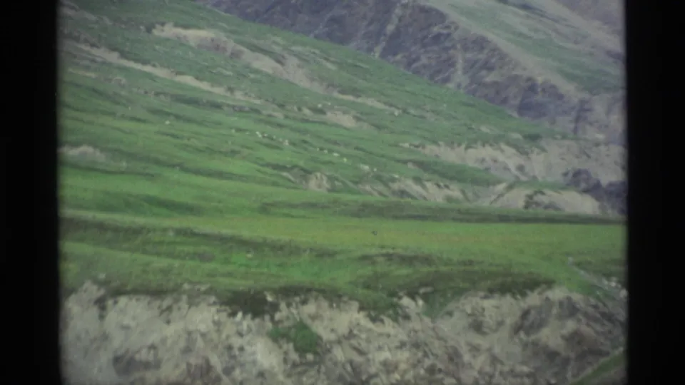 1977 Alaska 8mm frame emphasizing green mountain slopes, pale eroded ground, and distant rocky terrain.