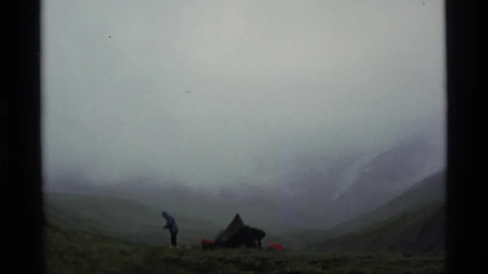 1977 Alaska 8mm frame with a distant adult in a blue jacket standing near a small tent in a foggy mountain valley.