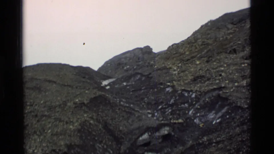 1977 Alaska 8mm frame showing rugged rocky mountain terrain, dark ridges, small snow patches, and a pale overcast sky.