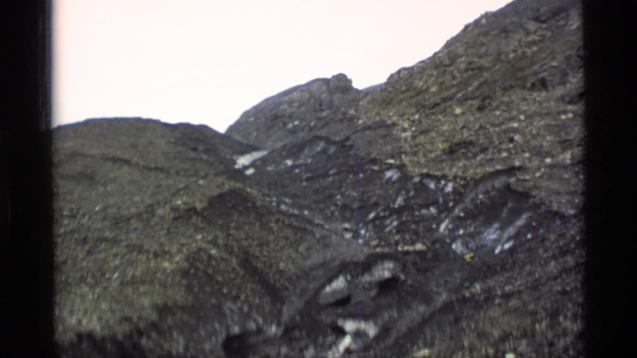 1977 Alaska 8mm frame showing steep rocky mountain terrain with scattered snow patches under a pale sky.