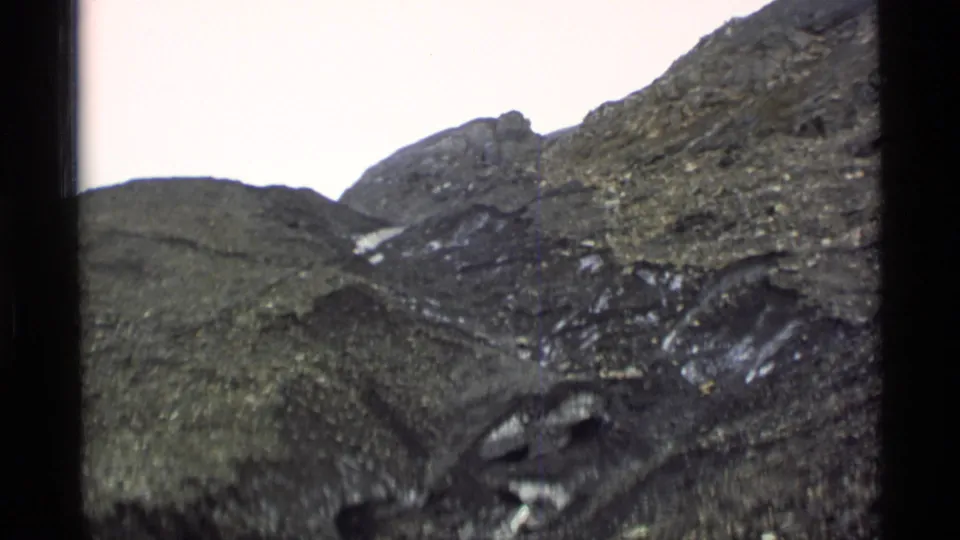 1977 Alaska 8mm frame showing steep rocky mountain terrain with scattered snow patches under a pale sky.