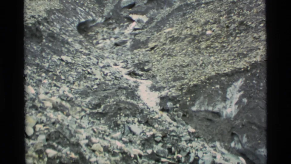 1977 Alaska 8mm frame showing a steep scree-covered mountainside with irregular snow streaks and dark rock gullies.