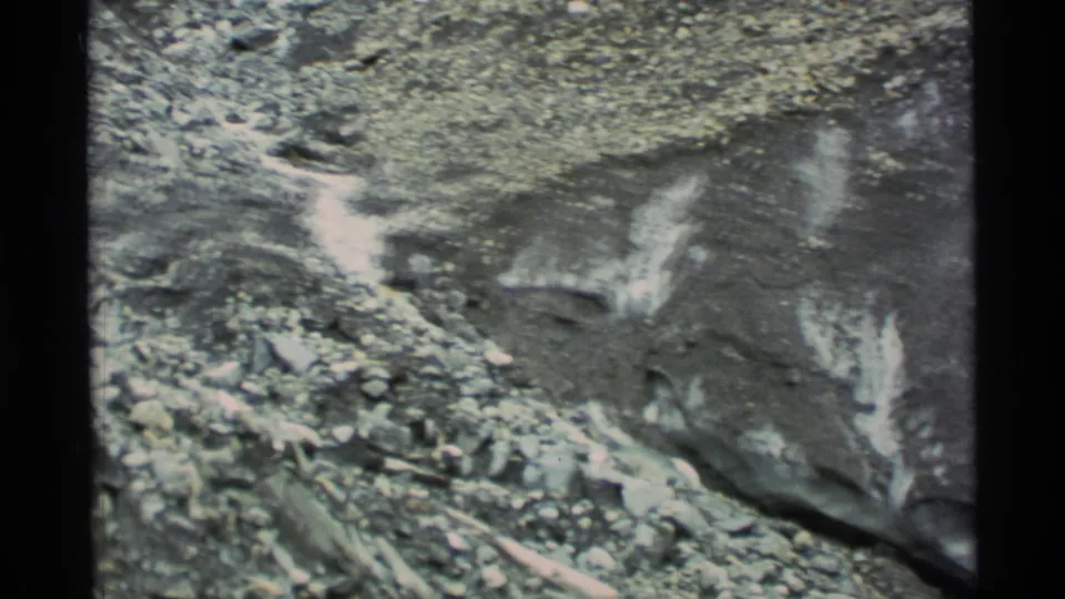 1977 Alaska 8mm frame with rocky mountain terrain, scattered snow patches, and a dark edge from the film or camera gate.