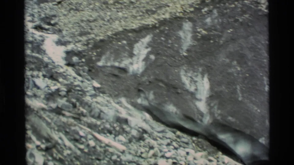 1977 Alaska 8mm frame showing a close rocky surface with pale mineral or ice-like streaks and scattered stones at the base.
