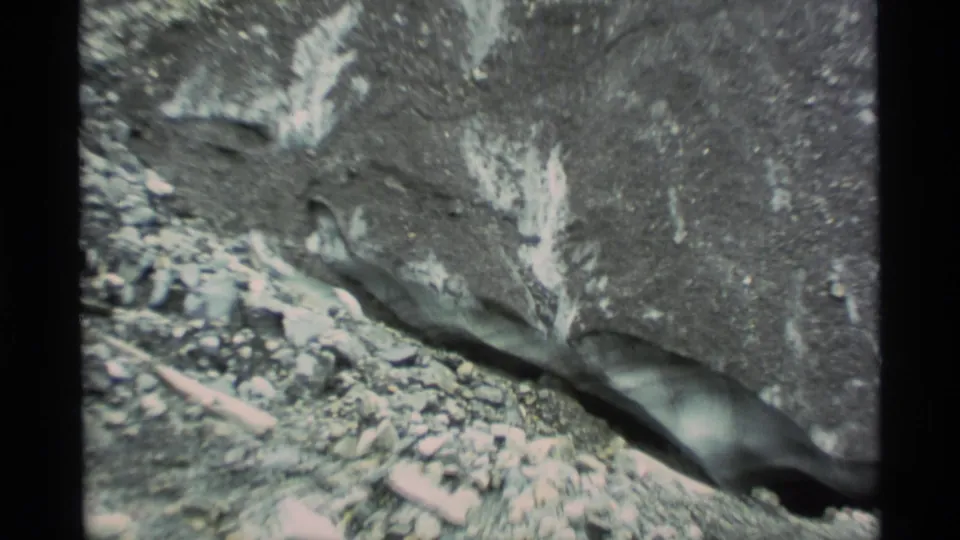 1977 Alaska 8mm frame of a dark rock face with pale streaks, a shadowed opening, and rocky debris in the foreground.