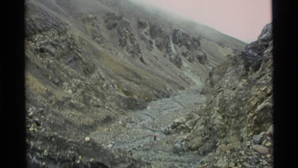 1977 Alaska 8mm frame showing a wide rugged valley, steep barren slopes, a rocky drainage bed, and a distant hiker in red.