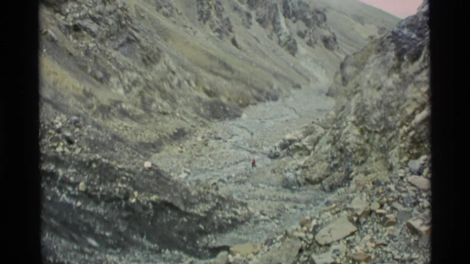 1977 Alaska 8mm frame looking down a rugged mountain valley with steep slopes, rocky foreground, and a pale drainage channel.
