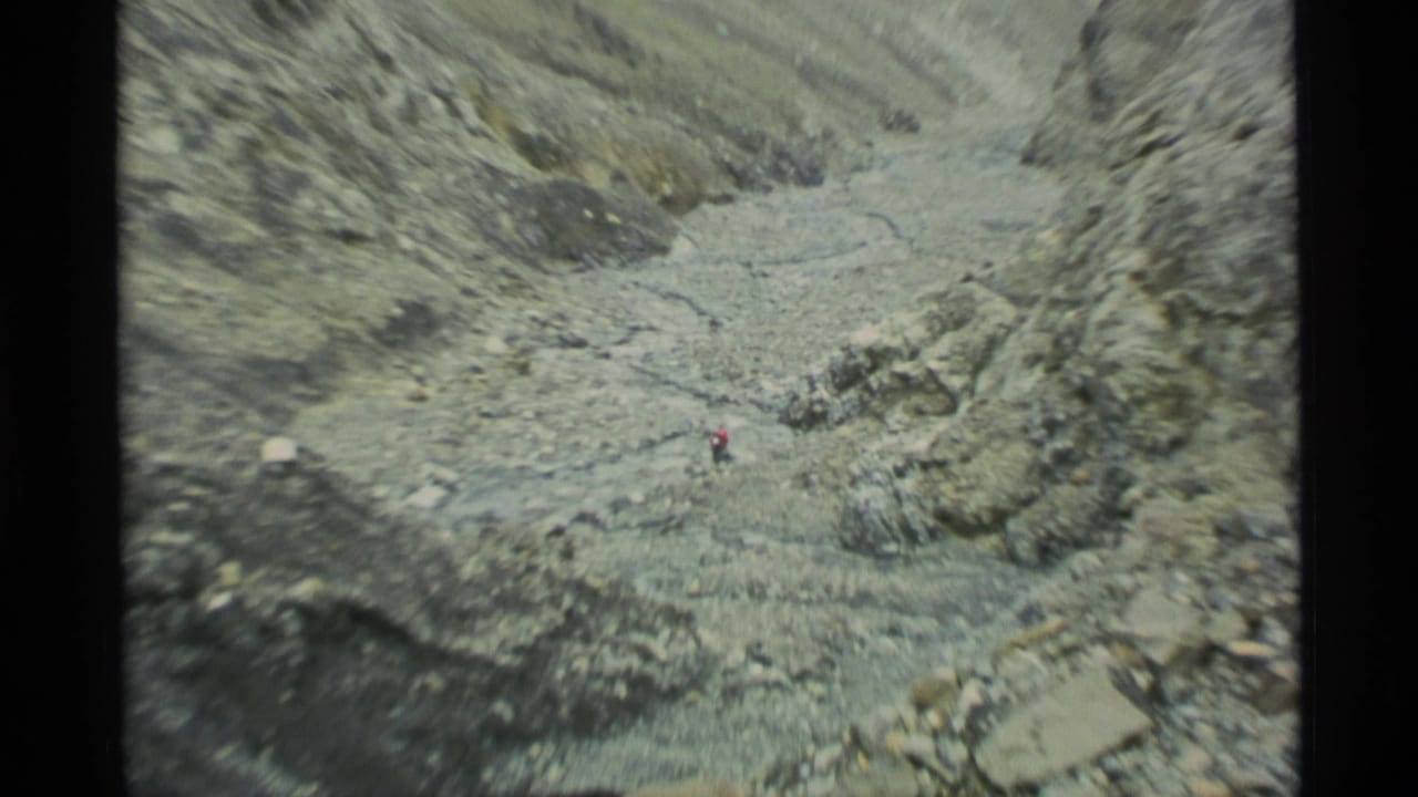 1977 Alaska 8mm frame showing a distant hiker moving through a barren rocky pass with loose scree and a pale stream channel.