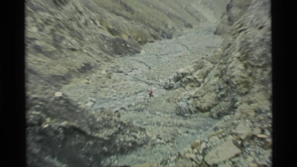1977 Alaska 8mm frame showing a distant hiker moving through a barren rocky pass with loose scree and a pale stream channel.