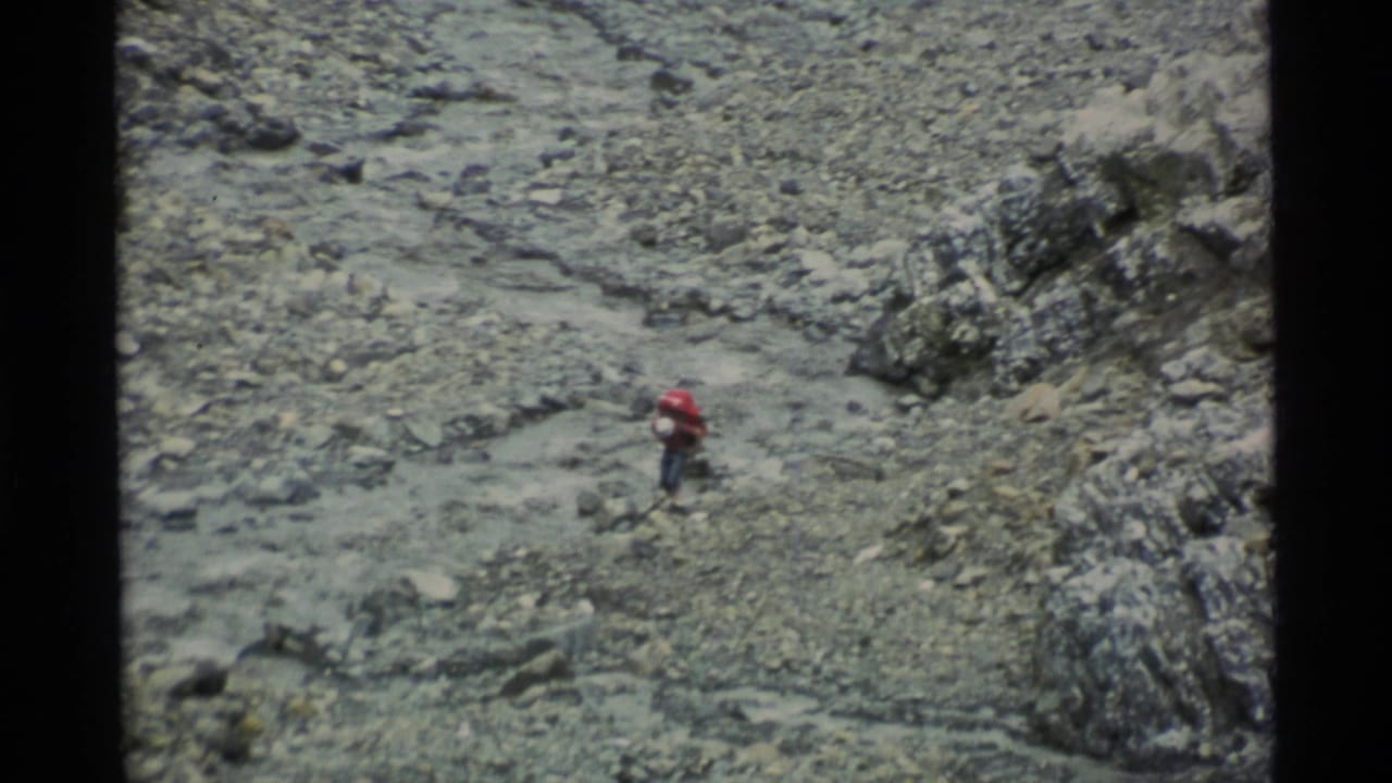 Hiker with a red pack walking across loose stones near rock outcrops.