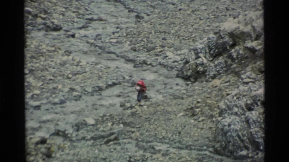 1977 Alaska home-movie frame of a hiker with a red pack navigating loose stones beside a narrow wash and dark rock outcrops.