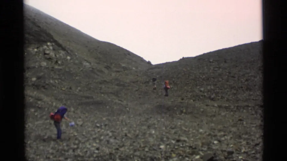 1977 Alaska 8mm frame with hikers spread across a barren slope, including one nearer figure with a purple pack and others ahead on the pass.