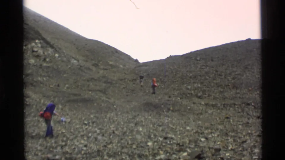1977 Alaska home-movie frame showing hikers climbing a broad rocky pass, with a nearer figure in a purple pack and others farther ahead.
