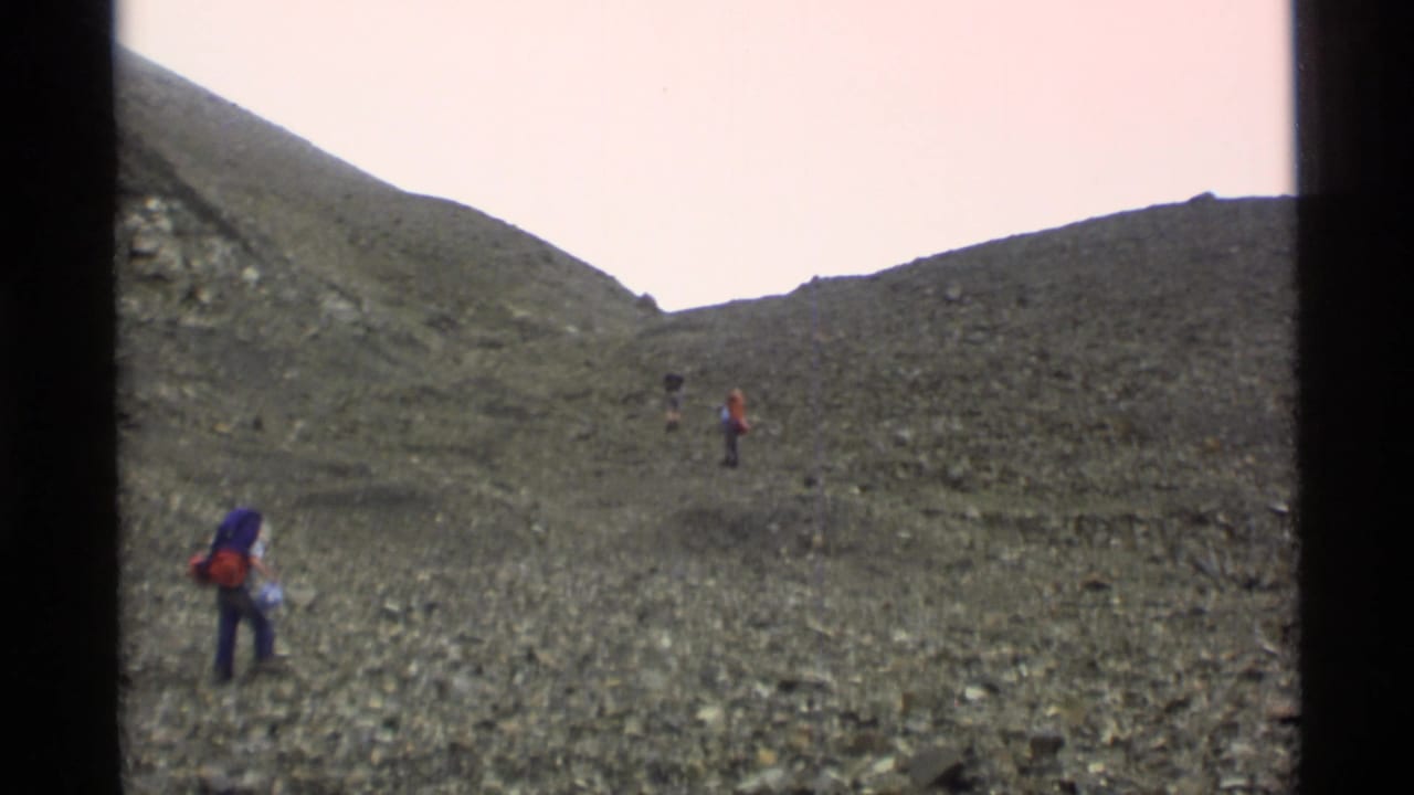 Backpackers cross a rocky mountain slope beneath a pale sky.
