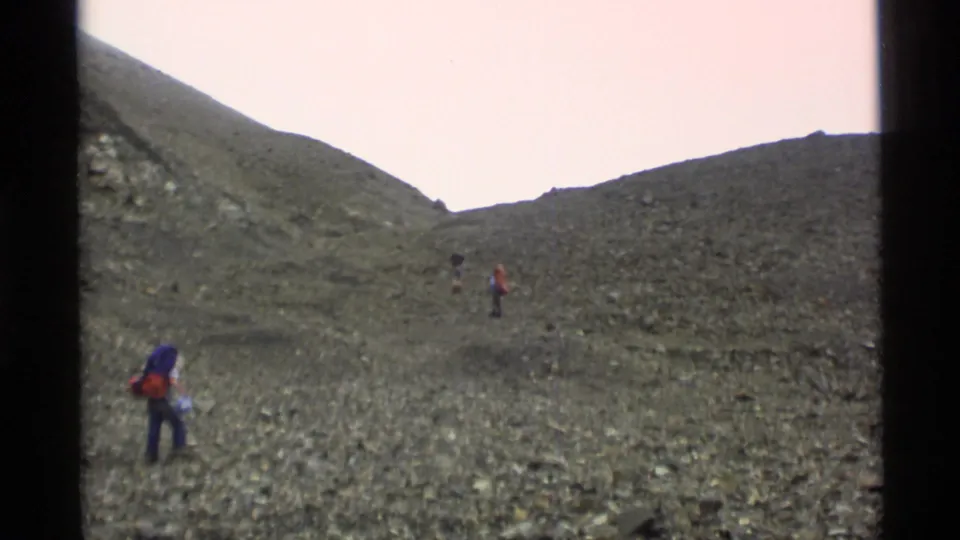 1977 Alaska home-movie frame showing backpacked hikers spread across a rocky mountain slope under a pale sky.