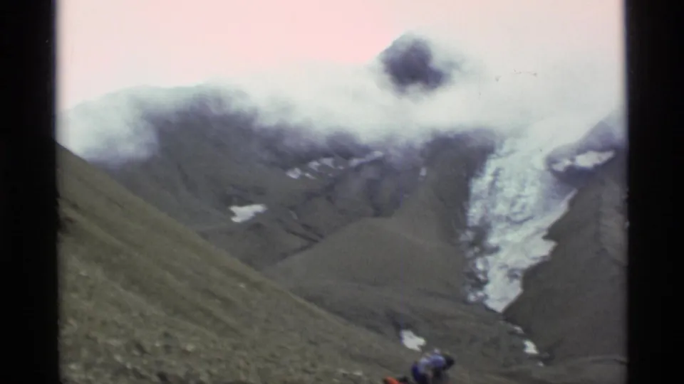 1977 Alaska frame showing a glacier below low clouds, with a small backpacked figure near the rocky foreground.