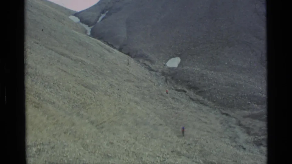 1977 Alaska 8mm frame showing tiny climbers on a broad green-gray mountain slope below dark ridges and snow patches.