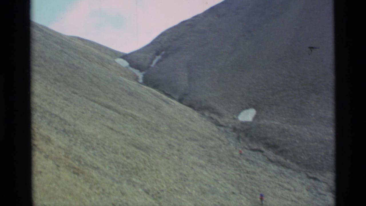 1977 Alaska home-movie frame of a broad mountain slope with snow remnants and a small climber near the bottom edge.