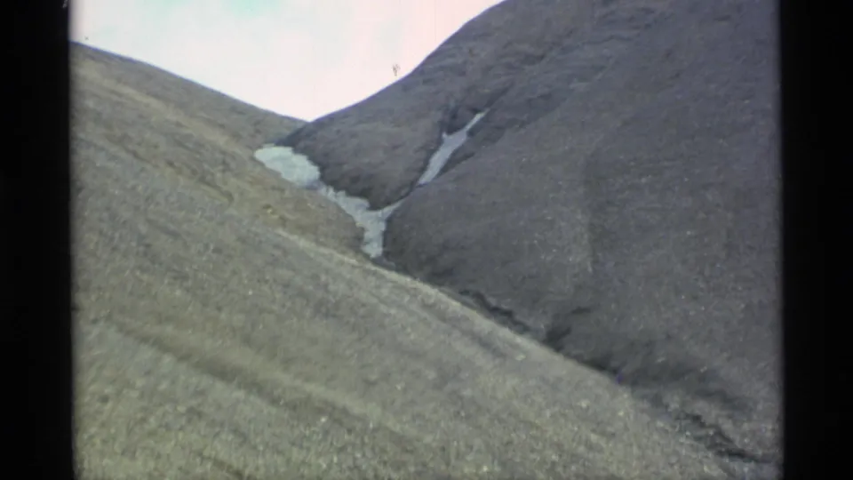 1977 Alaska home-movie frame showing a steep mountain slope, central snow patch, dark ridge, and soft 8mm color shifts.