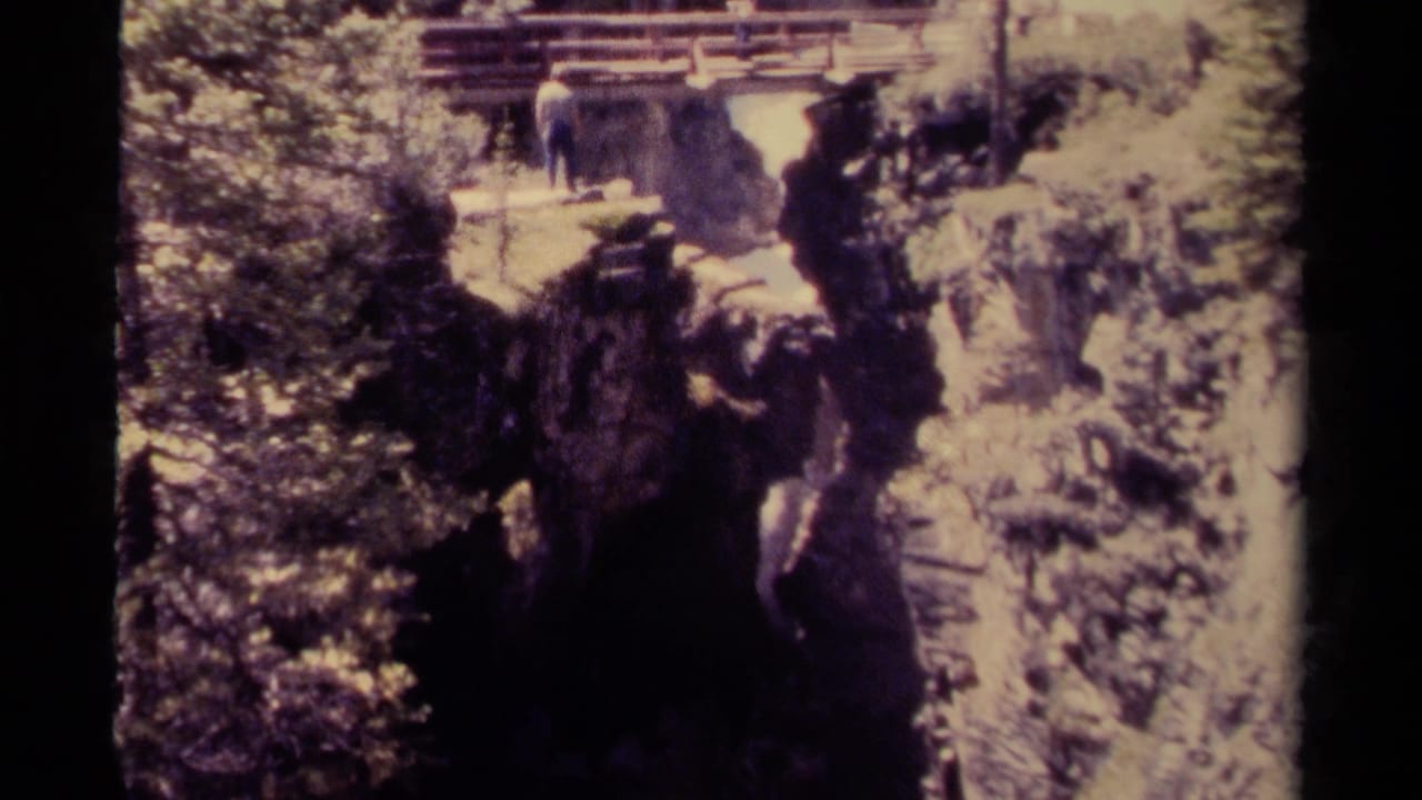The man remains near the wooden bridge above a shadowed gorge, with forest and pale rock surrounding the waterfall area.
