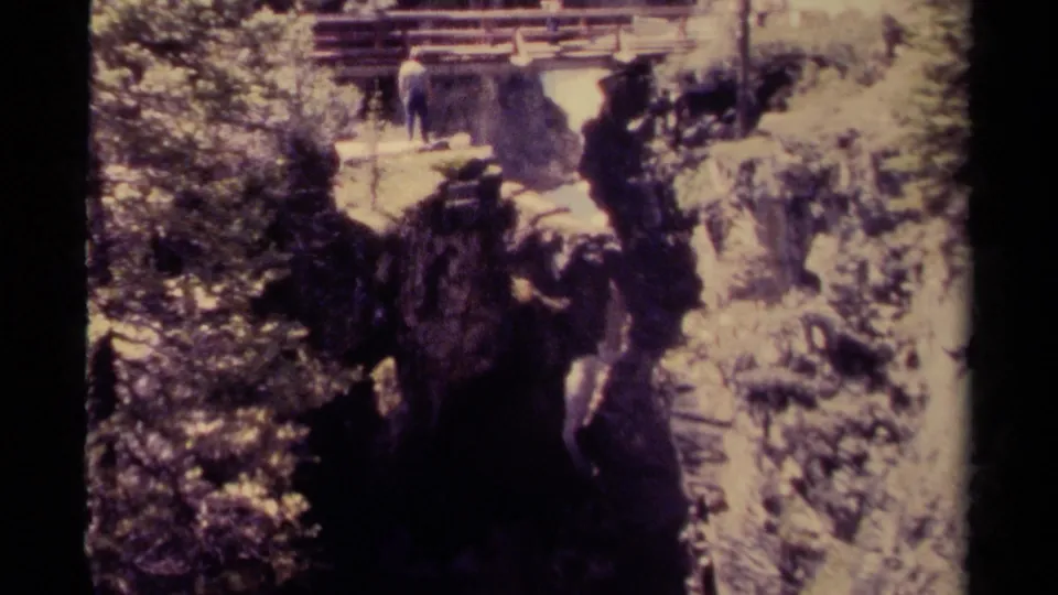 The man remains near the wooden bridge above a shadowed gorge, with forest and pale rock surrounding the waterfall area.