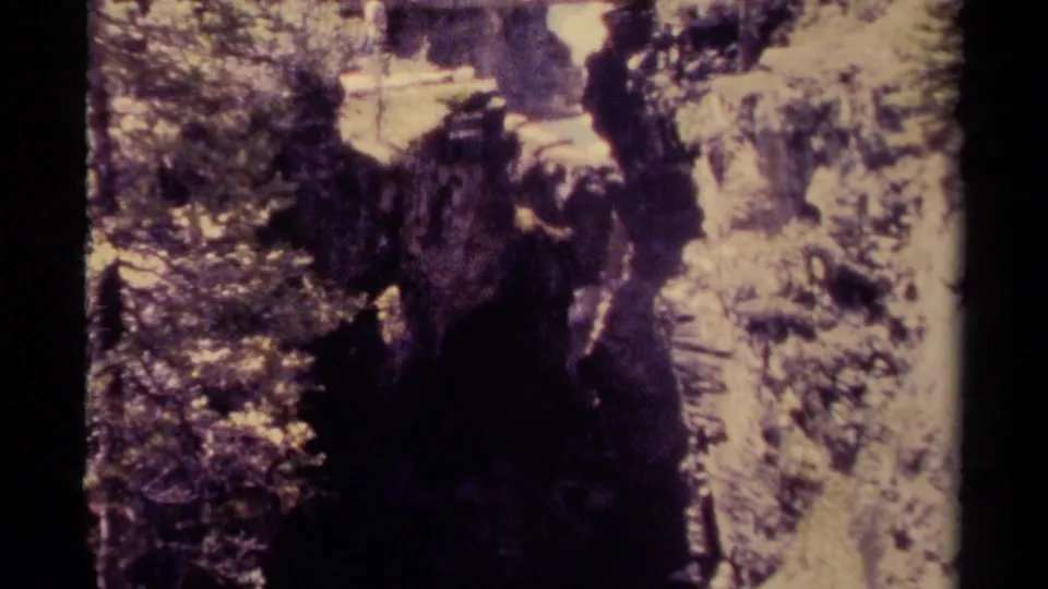 The handheld 8mm view shifts down the canyon, showing dark rock, foliage, and water with the bridge partly out of frame.
