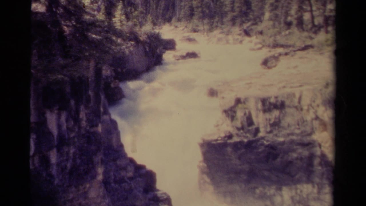1979 Canada 8mm frame of a rushing river or waterfall cutting through steep dark rock with forest visible along the upper bank.