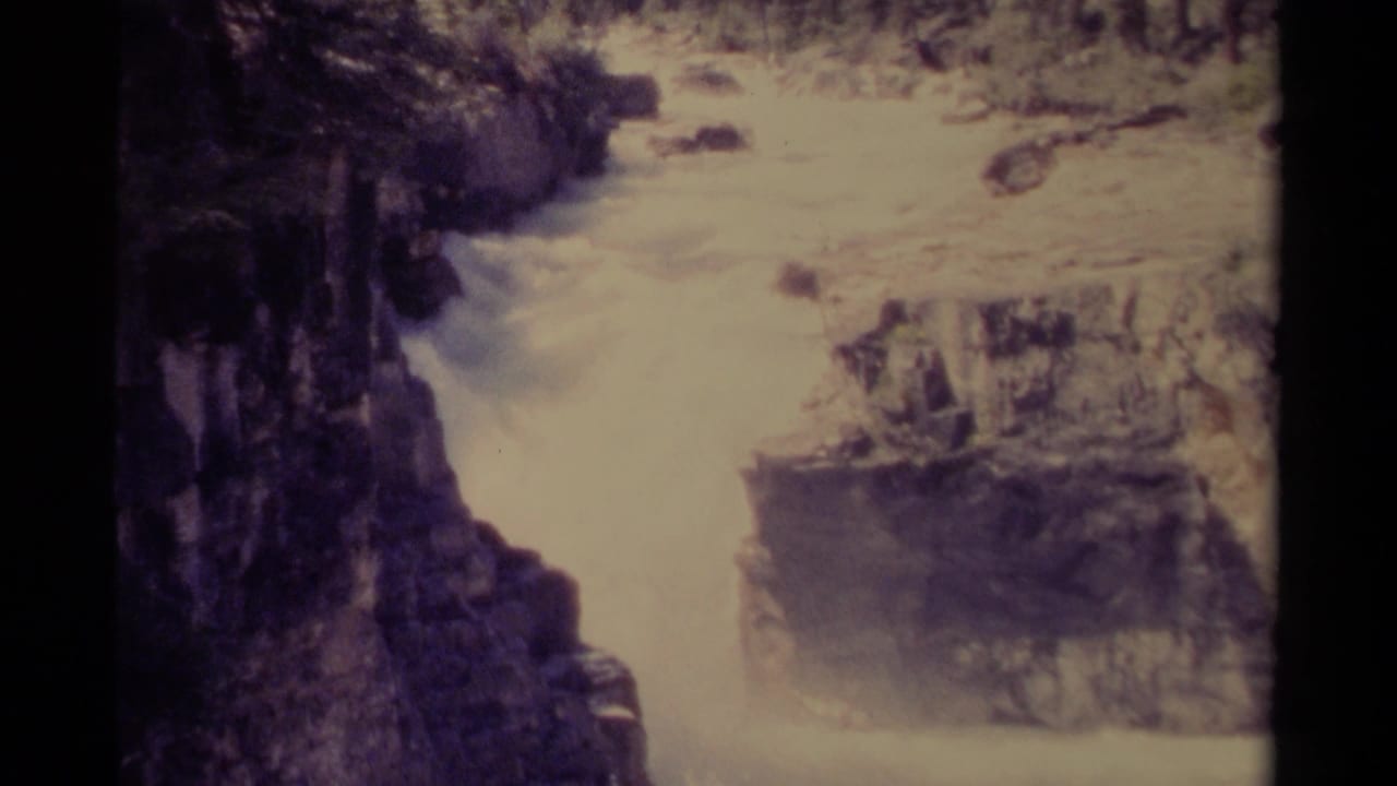 Archival 8mm view in Canada of turbulent whitewater moving through a rocky mountain gorge with trees along the top edge.